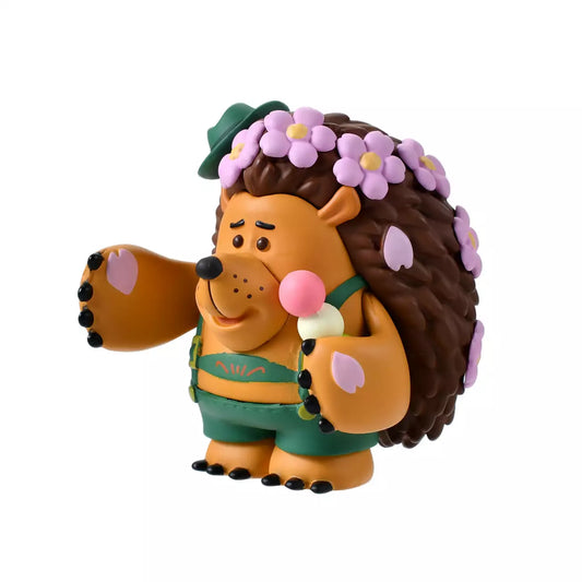 A figure of Mr. Pricklepants from Toy Story 3 enjoying a spring picnic. The hedgehog wears green lederhosen and a small green hat. He holds a 'Hanami Dango' (a three-colored sweet dumpling) in one hand and has a crown of pink cherry blossoms on his head. The background is pink with falling sakura petals.
