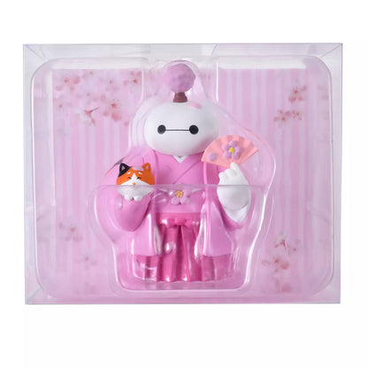 An adorable figure of Baymax from Big Hero 6 dressed in a traditional pink kimono and hakama for the cherry blossom season. Baymax holds a pink fan with a flower design in one hand and cradles Mochi the cat (also smiling) in the other arm. A small cherry blossom tree sprouts from Baymax's head. The setting is a festive pink sakura background.