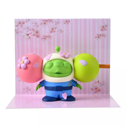 A funny and creative figure of a Little Green Man (Alien) reimagined as a 'Hanami Dango' skewer. The Alien's head is the center green dumpling, sandwiched between a pink dumpling on the left and a lime green dumpling on the right. The character wears a blue space suit body below the dango head. Pink flower petals decorate the dumplings.