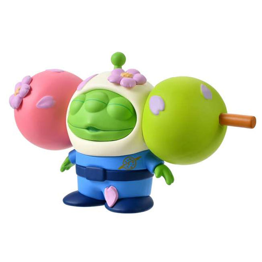 A funny and creative figure of a Little Green Man (Alien) reimagined as a 'Hanami Dango' skewer. The Alien's head is the center green dumpling, sandwiched between a pink dumpling on the left and a lime green dumpling on the right. The character wears a blue space suit body below the dango head. Pink flower petals decorate the dumplings.