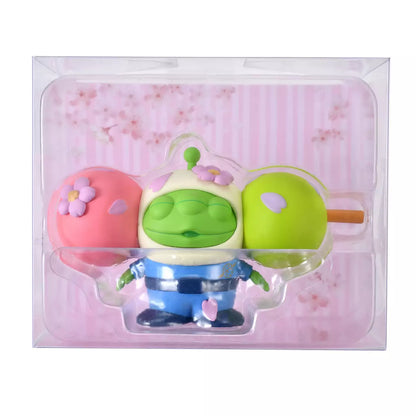 A funny and creative figure of a Little Green Man (Alien) reimagined as a 'Hanami Dango' skewer. The Alien's head is the center green dumpling, sandwiched between a pink dumpling on the left and a lime green dumpling on the right. The character wears a blue space suit body below the dango head. Pink flower petals decorate the dumplings.
