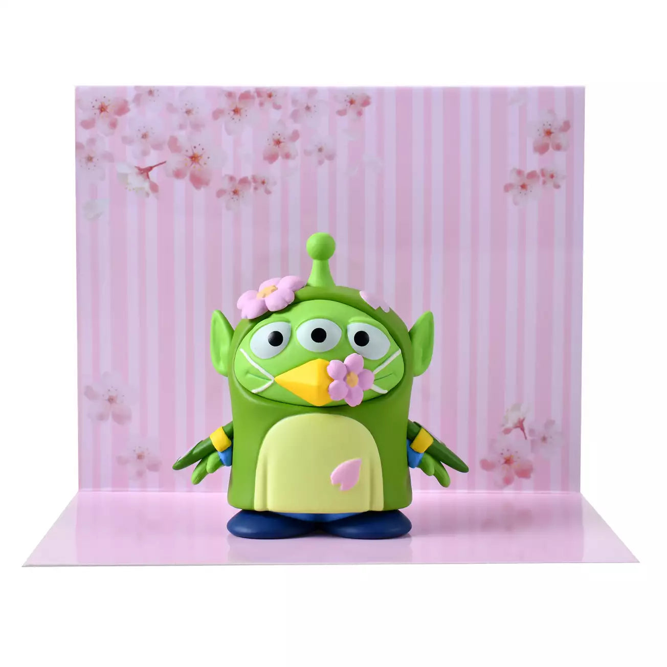 A whimsical figure of a Little Green Man (Alien) disguised as a Japanese Bush Warbler (Uguisu). The Alien is wearing a green bird costume with yellow beak details and blue accents on the wings. Pink cherry blossom flowers decorate the costume's head and beak. The figure stands in front of a scenic pink background with sakura motifs.