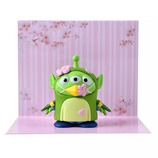 A whimsical figure of a Little Green Man (Alien) disguised as a Japanese Bush Warbler (Uguisu). The Alien is wearing a green bird costume with yellow beak details and blue accents on the wings. Pink cherry blossom flowers decorate the costume's head and beak. The figure stands in front of a scenic pink background with sakura motifs.