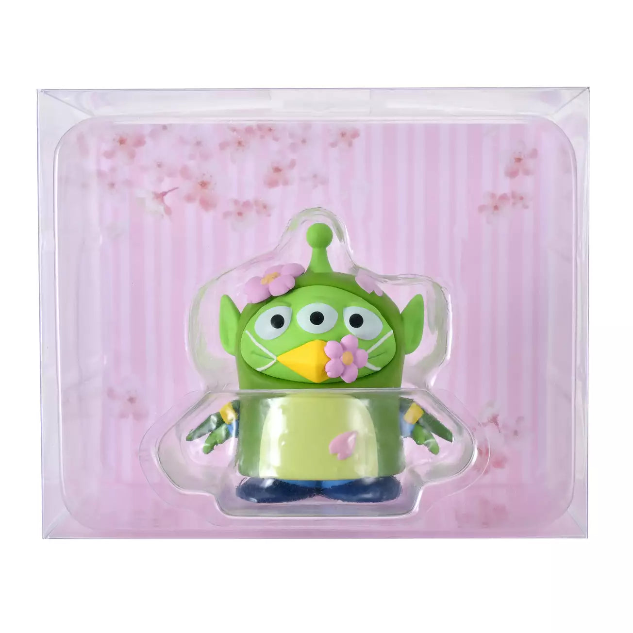 A whimsical figure of a Little Green Man (Alien) disguised as a Japanese Bush Warbler (Uguisu). The Alien is wearing a green bird costume with yellow beak details and blue accents on the wings. Pink cherry blossom flowers decorate the costume's head and beak. The figure stands in front of a scenic pink background with sakura motifs.