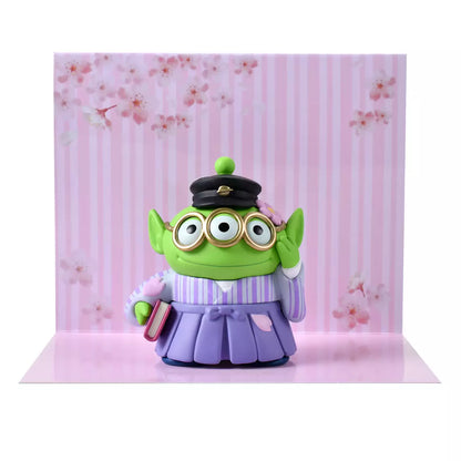A Disney Store Japan exclusive figure of a Little Green Man (Alien) from Toy Story. The character is dressed in a traditional Japanese purple striped hakama and wears a black student cap. It poses with a book in one hand and touches its glasses with the other. The figure is set against a pink striped background with falling cherry blossom petals.