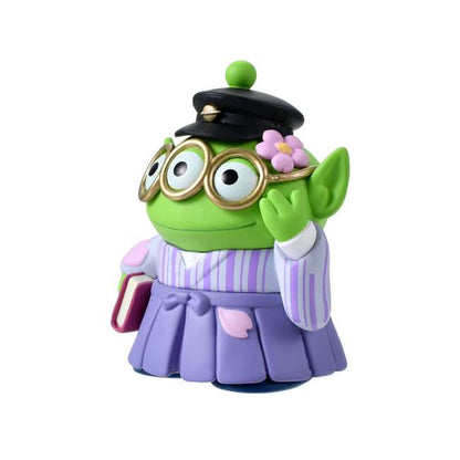 A Disney Store Japan exclusive figure of a Little Green Man (Alien) from Toy Story. The character is dressed in a traditional Japanese purple striped hakama and wears a black student cap. It poses with a book in one hand and touches its glasses with the other. The figure is set against a pink striped background with falling cherry blossom petals.