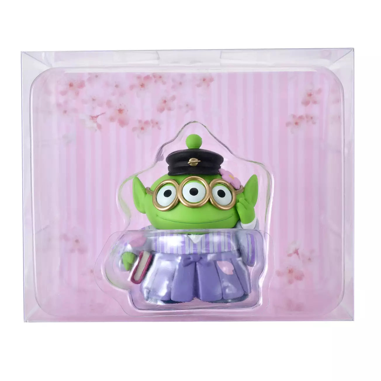A Disney Store Japan exclusive figure of a Little Green Man (Alien) from Toy Story. The character is dressed in a traditional Japanese purple striped hakama and wears a black student cap. It poses with a book in one hand and touches its glasses with the other. The figure is set against a pink striped background with falling cherry blossom petals.