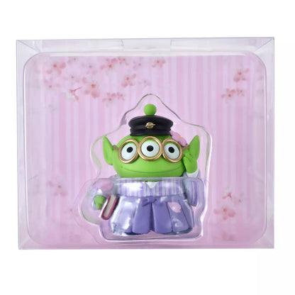 A Disney Store Japan exclusive figure of a Little Green Man (Alien) from Toy Story. The character is dressed in a traditional Japanese purple striped hakama and wears a black student cap. It poses with a book in one hand and touches its glasses with the other. The figure is set against a pink striped background with falling cherry blossom petals.