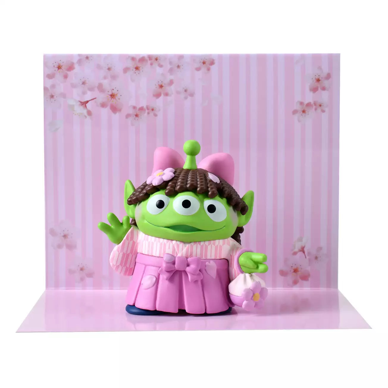 A cute collectible figure of a Little Green Man (Alien) wearing a pink wig with brown pigtails and a traditional pink hakama kimono. The character holds a small flower-shaped purse and waves with one hand. The backdrop features a pink striped pattern with delicate cherry blossom illustrations.