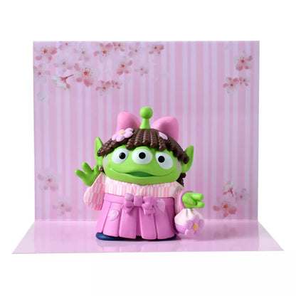 A cute collectible figure of a Little Green Man (Alien) wearing a pink wig with brown pigtails and a traditional pink hakama kimono. The character holds a small flower-shaped purse and waves with one hand. The backdrop features a pink striped pattern with delicate cherry blossom illustrations.