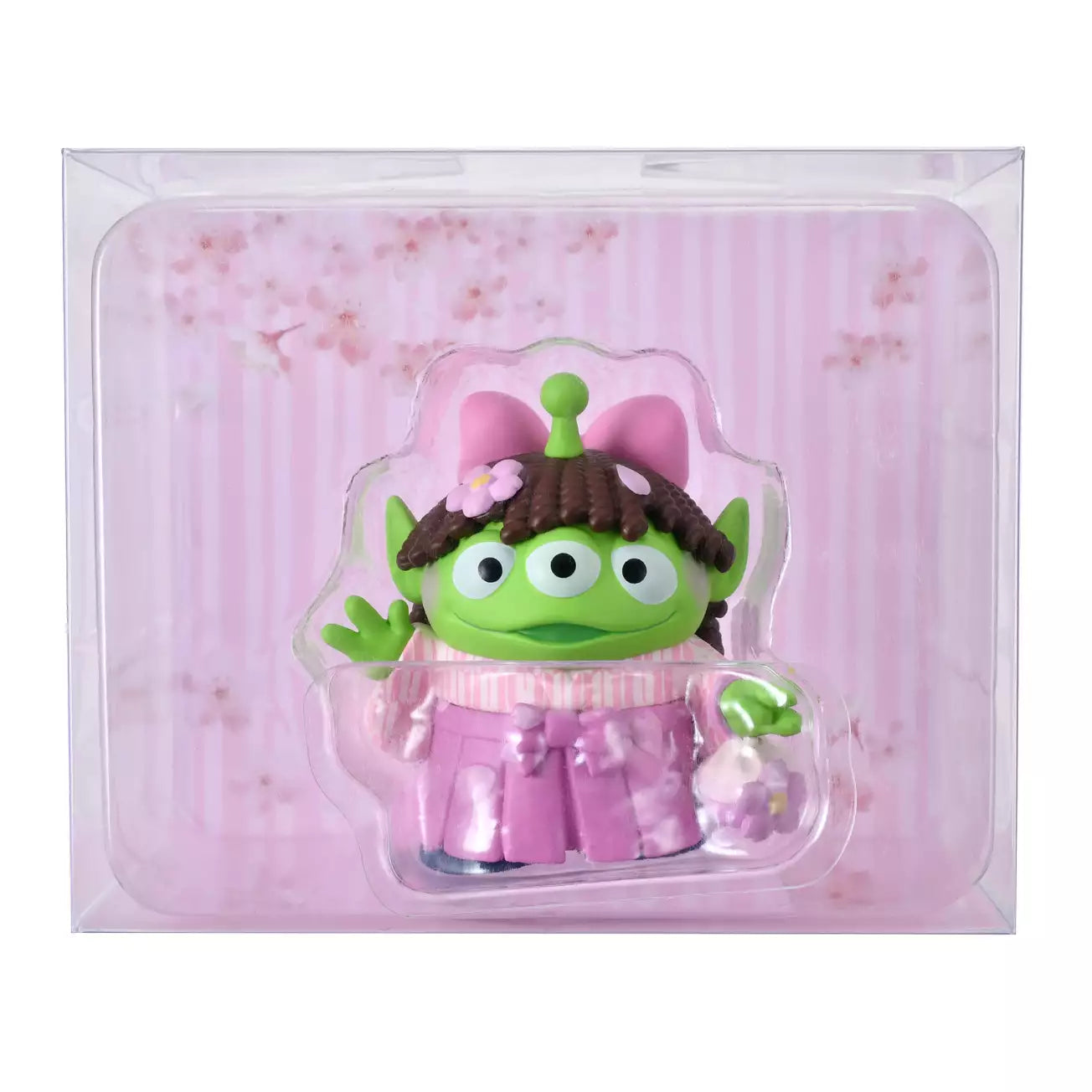 A cute collectible figure of a Little Green Man (Alien) wearing a pink wig with brown pigtails and a traditional pink hakama kimono. The character holds a small flower-shaped purse and waves with one hand. The backdrop features a pink striped pattern with delicate cherry blossom illustrations.