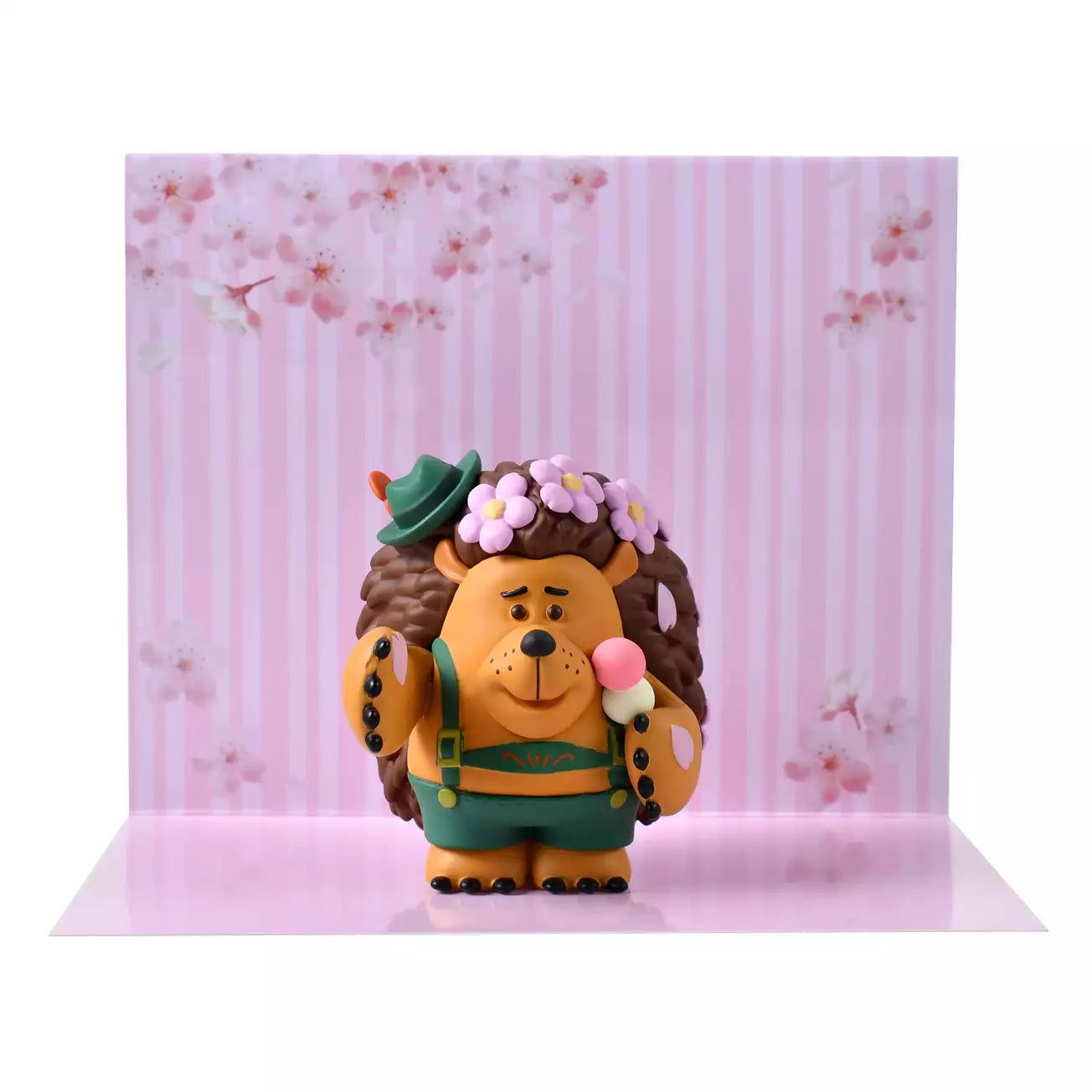 A figure of Mr. Pricklepants from Toy Story 3 enjoying a spring picnic. The hedgehog wears green lederhosen and a small green hat. He holds a 'Hanami Dango' (a three-colored sweet dumpling) in one hand and has a crown of pink cherry blossoms on his head. The background is pink with falling sakura petals.