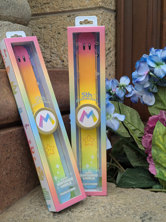 Two packaged limited-edition Super Nintendo World 5th Anniversary Power-Up Bands from Universal Studios Japan, featuring a pastel rainbow gradient strap, gold star details, and a gold-rimmed Mario 'M' emblem.
