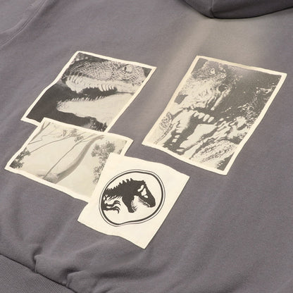 A grey zip-up hoodie from the USJ UNIVRS collection with a subtle graphic of a dinosaur scratching its claws and the text 'JURASSIC WORLD' on the front. The back features four vintage comic panels of dinosaurs.